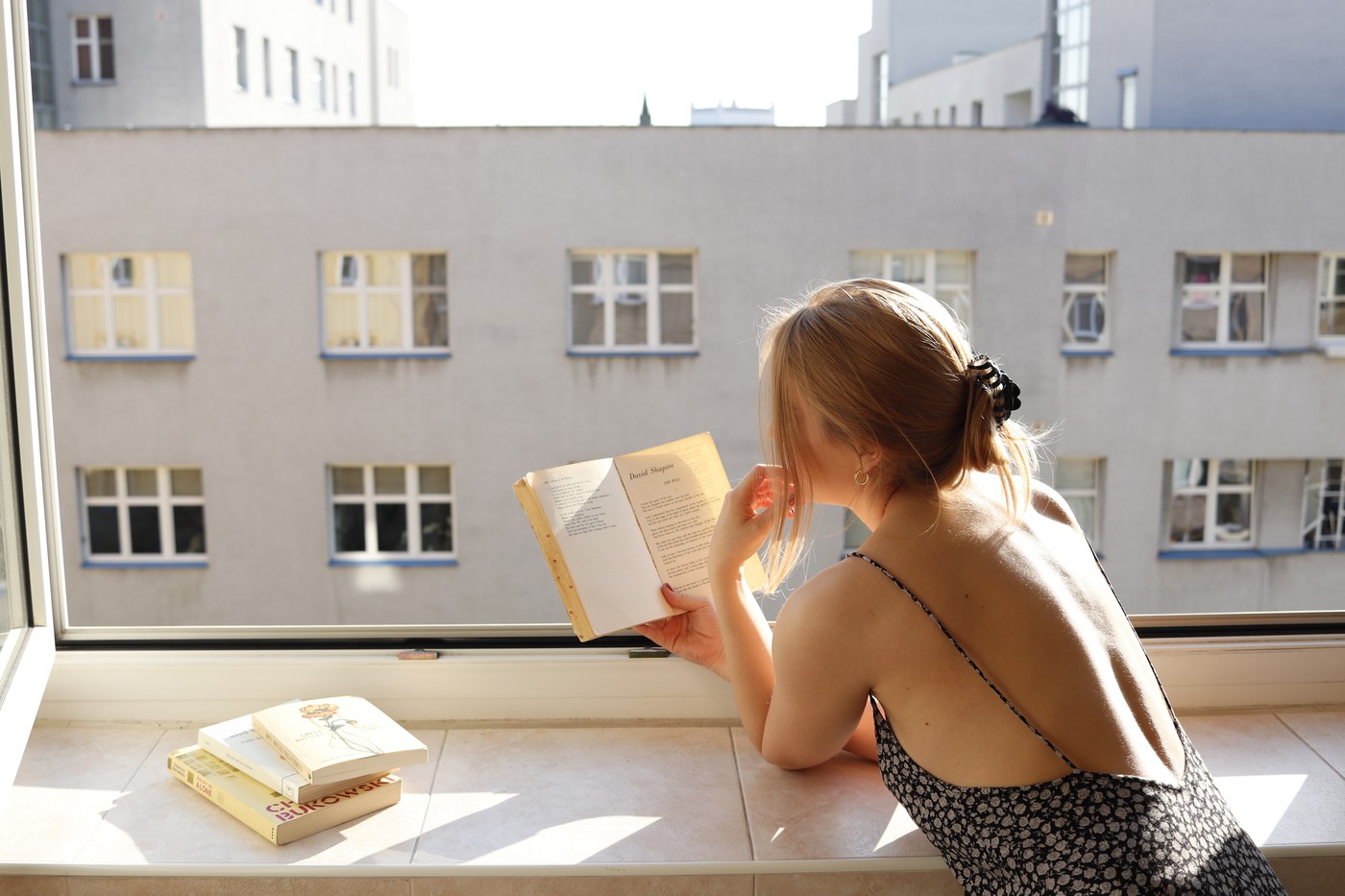 Anna reading by an open window, morning light, a small stack of books on the sill.