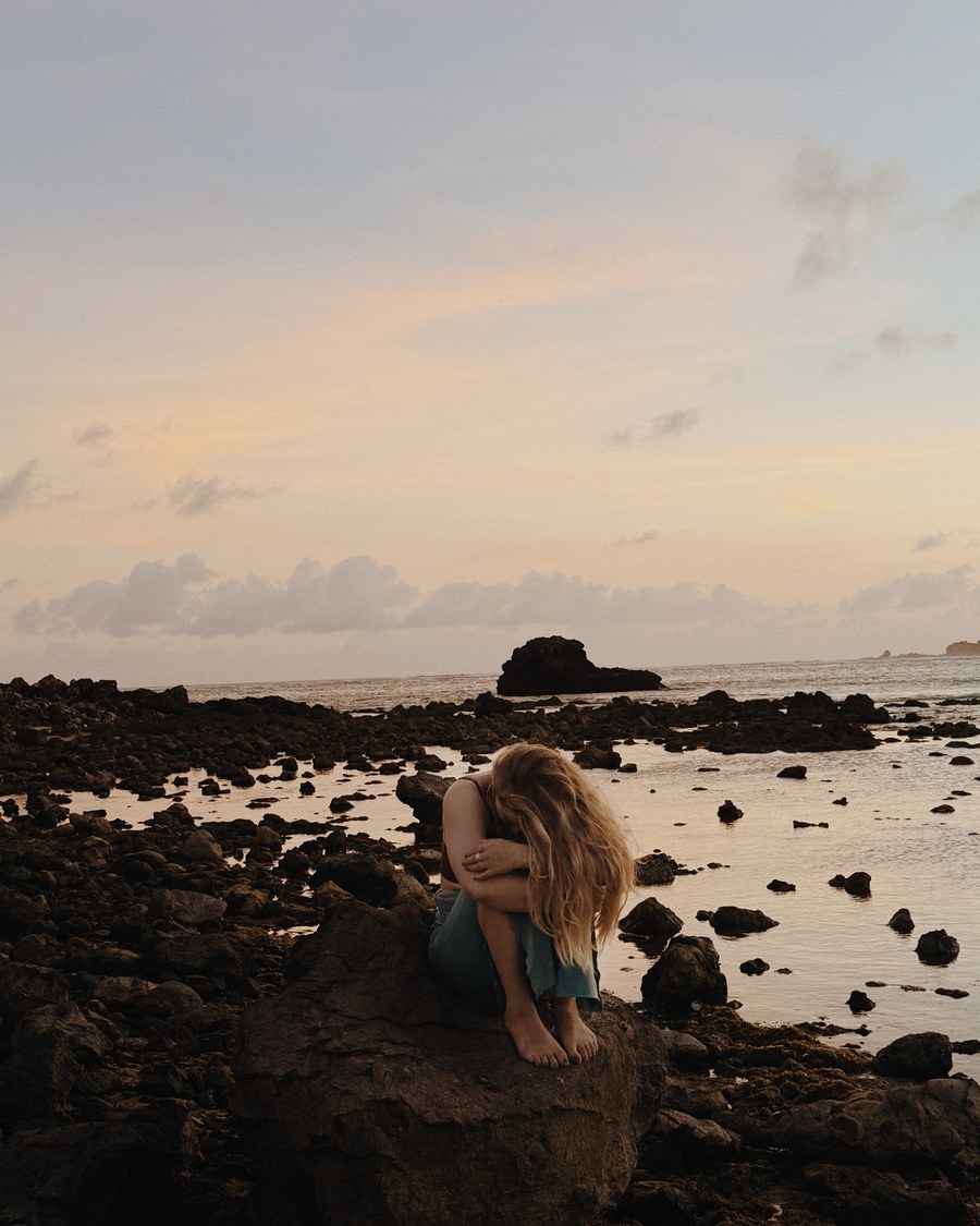 Anna sitting quietly on rocks by the sea at dusk, a reflective moment.